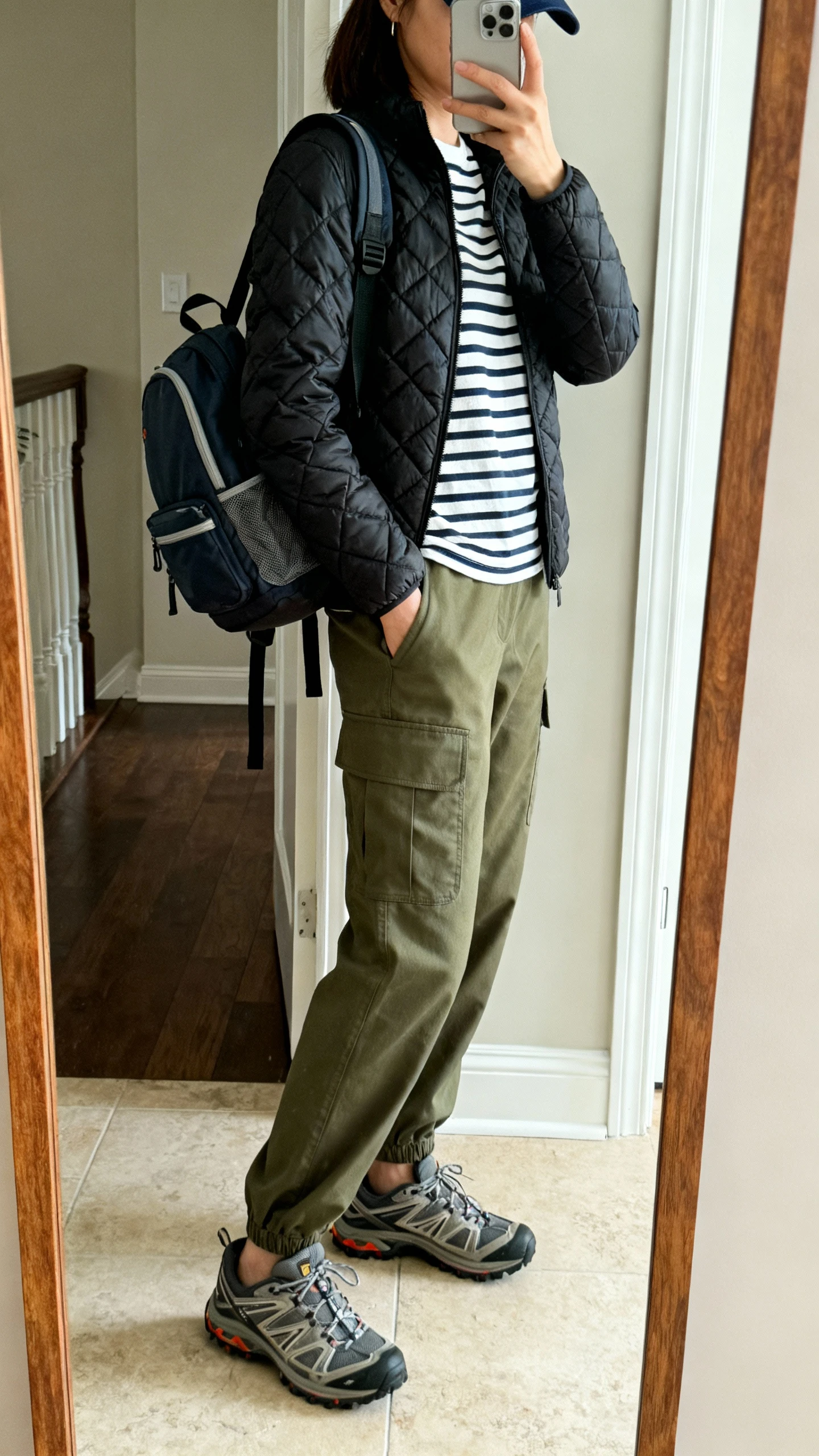 iPhone side-angle mirror selfie of a woman wearing a utility-chic combo: olive cargo pants, striped Breton tee, black quilted liner jacket, trail sneakers, compact backpack, baseball cap, face not visible, full-length mirror near entryway, natural daylight, iPhone photo quality.