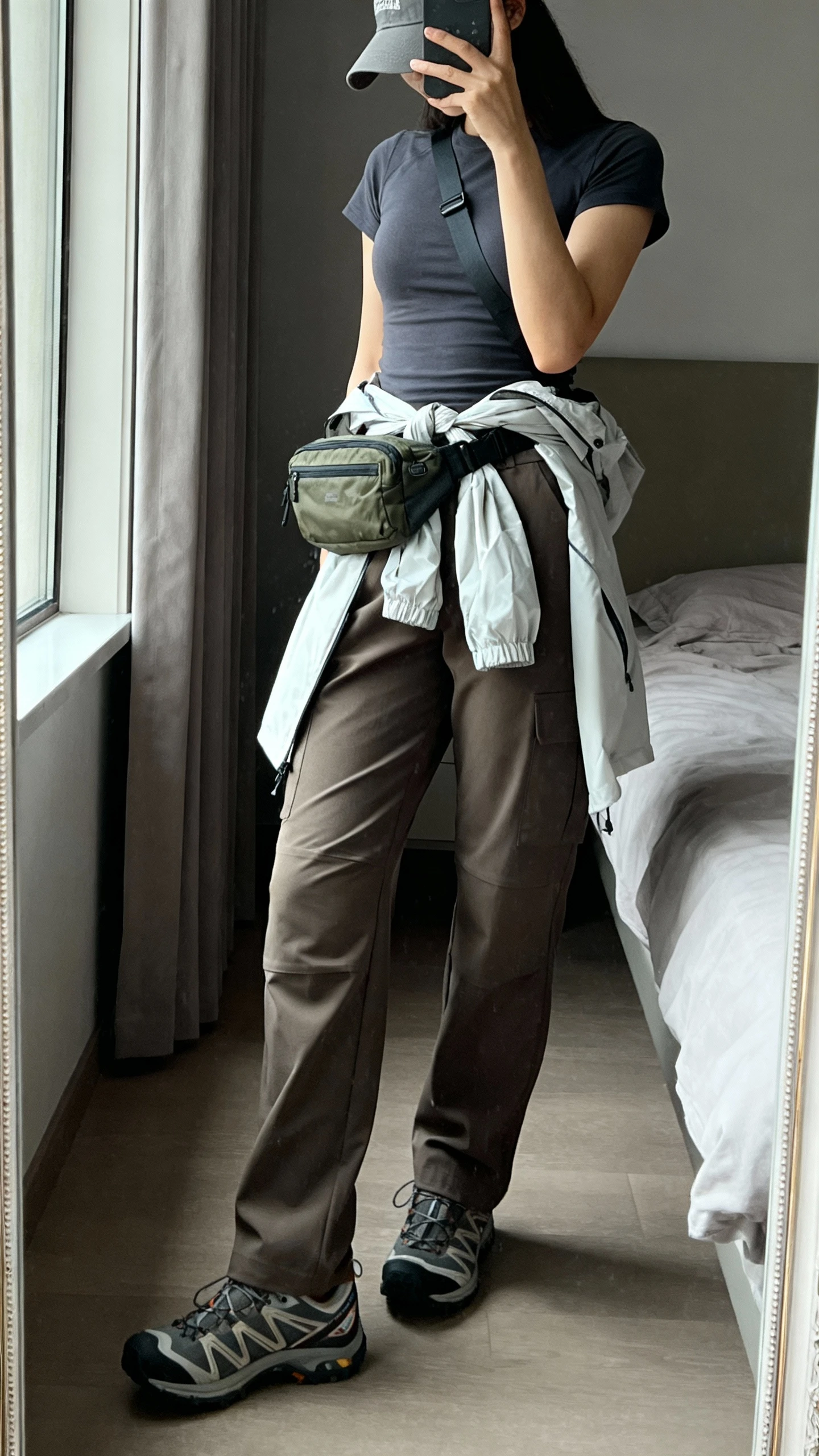 iPhone side-angle mirror selfie of a woman wearing tailored cargo pants, fitted crewneck tee, trail sneakers, nylon belt bag worn crossbody, lightweight packable windbreaker tied at waist, baseball cap, face not visible, bedroom mirror, natural window light, iPhone photo quality.