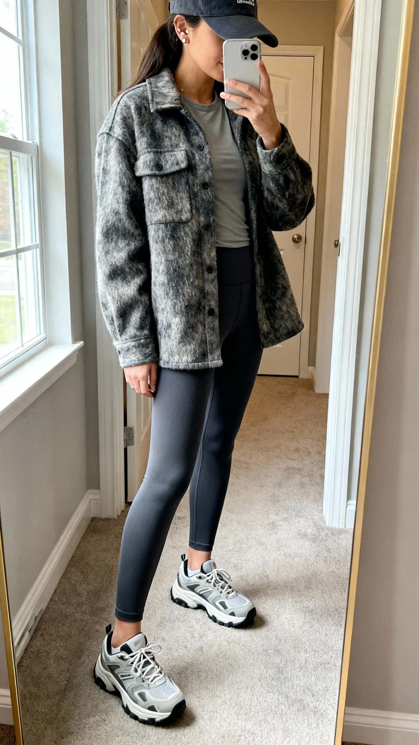 iPhone side-angle mirror selfie of a woman wearing a brushed shacket over a fitted tee with tailored leggings, chunky sneakers, baseball cap, and simple studs, face not visible, cropped at shoulders, entryway mirror, natural window light, iPhone photo quality.