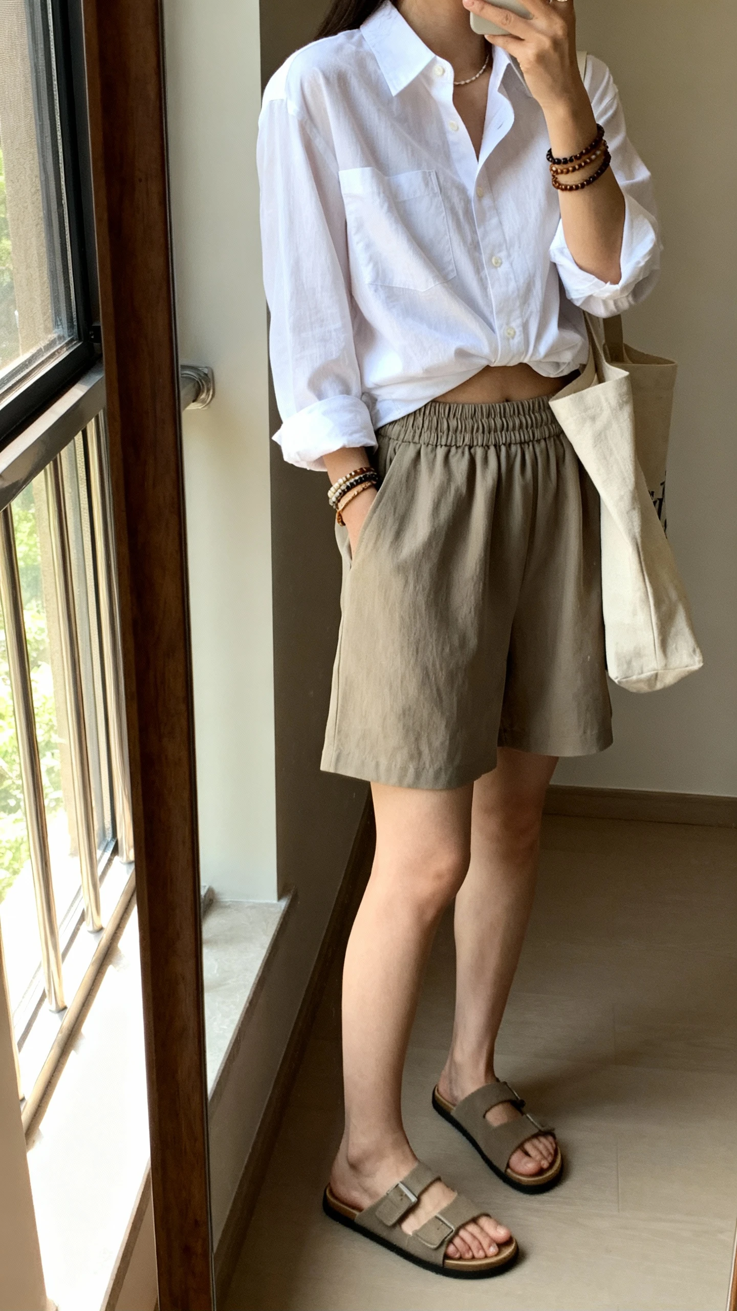 iPhone side-angle selfie of a woman wearing a crisp button-down shirt half-tucked into pull-on shorts with an elastic waist, slide sandals, layered bracelets, and a canvas tote, face not visible, full-length mirror near window, afternoon natural light, iPhone photo quality